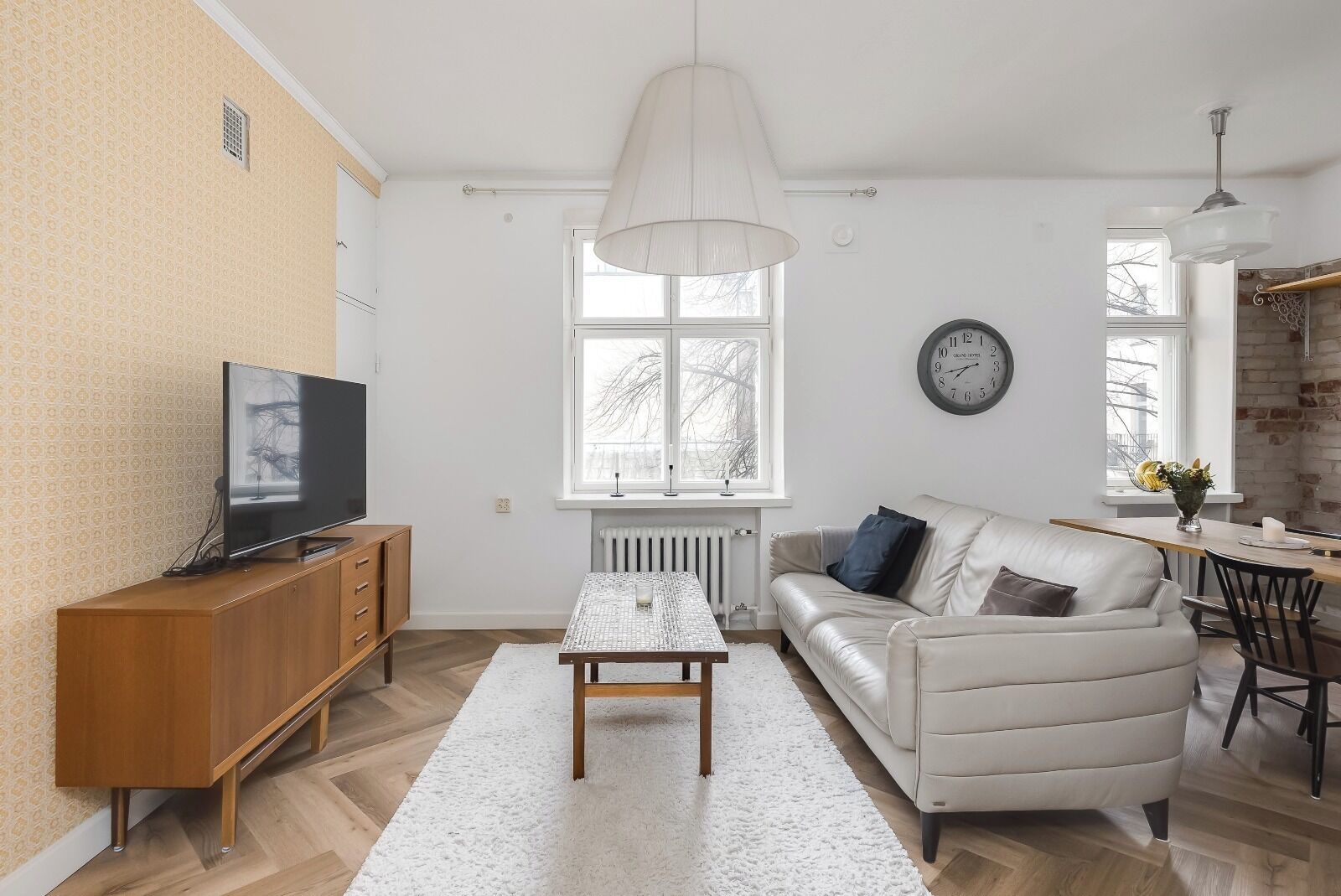A modern living room with a sofa, a coffee table, a TV on a wooden cabinet, a large window, and a clock on the wall.