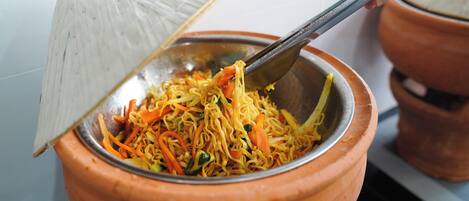 A hand using a metal utensil to serve noodles from a clay pot.
