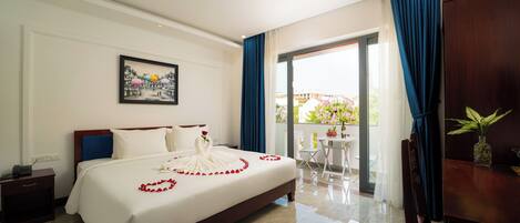 A hotel room with a large bed, a bedside table, a nightstand, a window with blue curtains, a balcony with a table and chairs, and a framed picture on the wall.