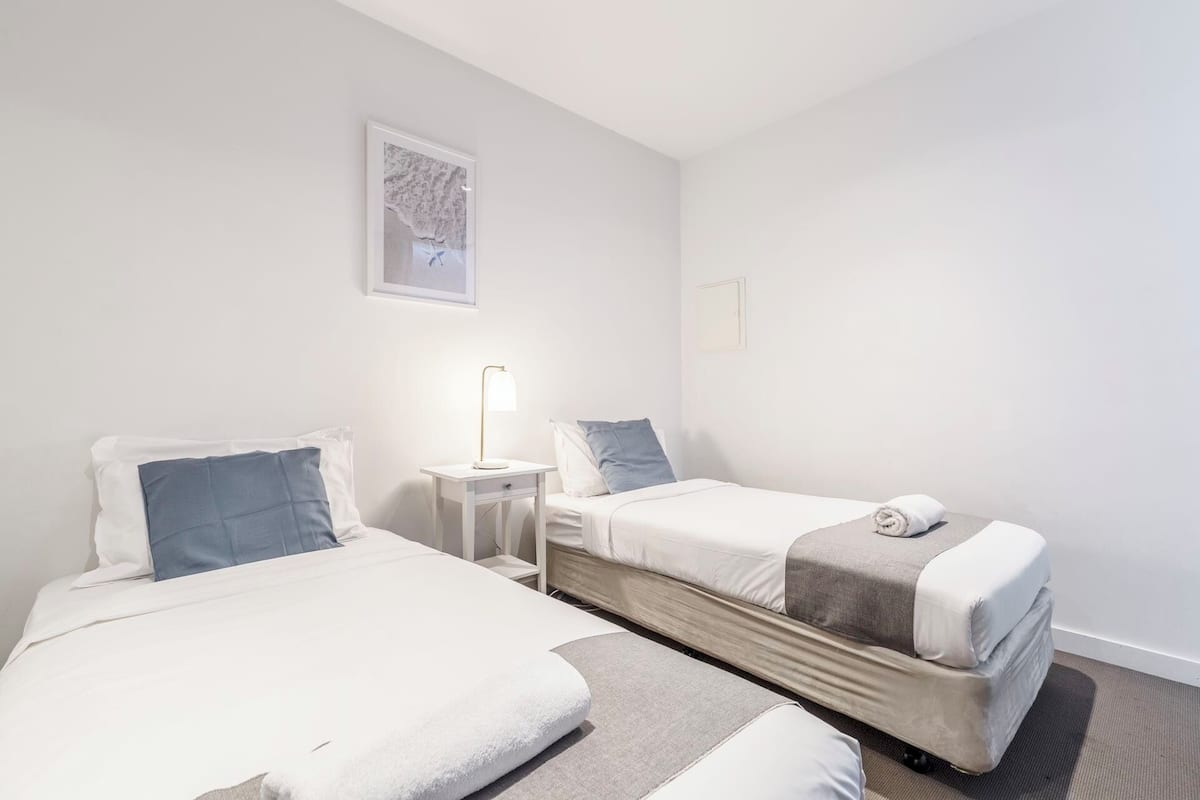 A hotel room with two beds, a bedside table with a lamp, and a framed picture on the wall.