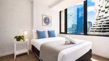 A modern hotel room with a large bed, a bedside table with a lamp, a framed artwork, and a view of a cityscape through a large window.