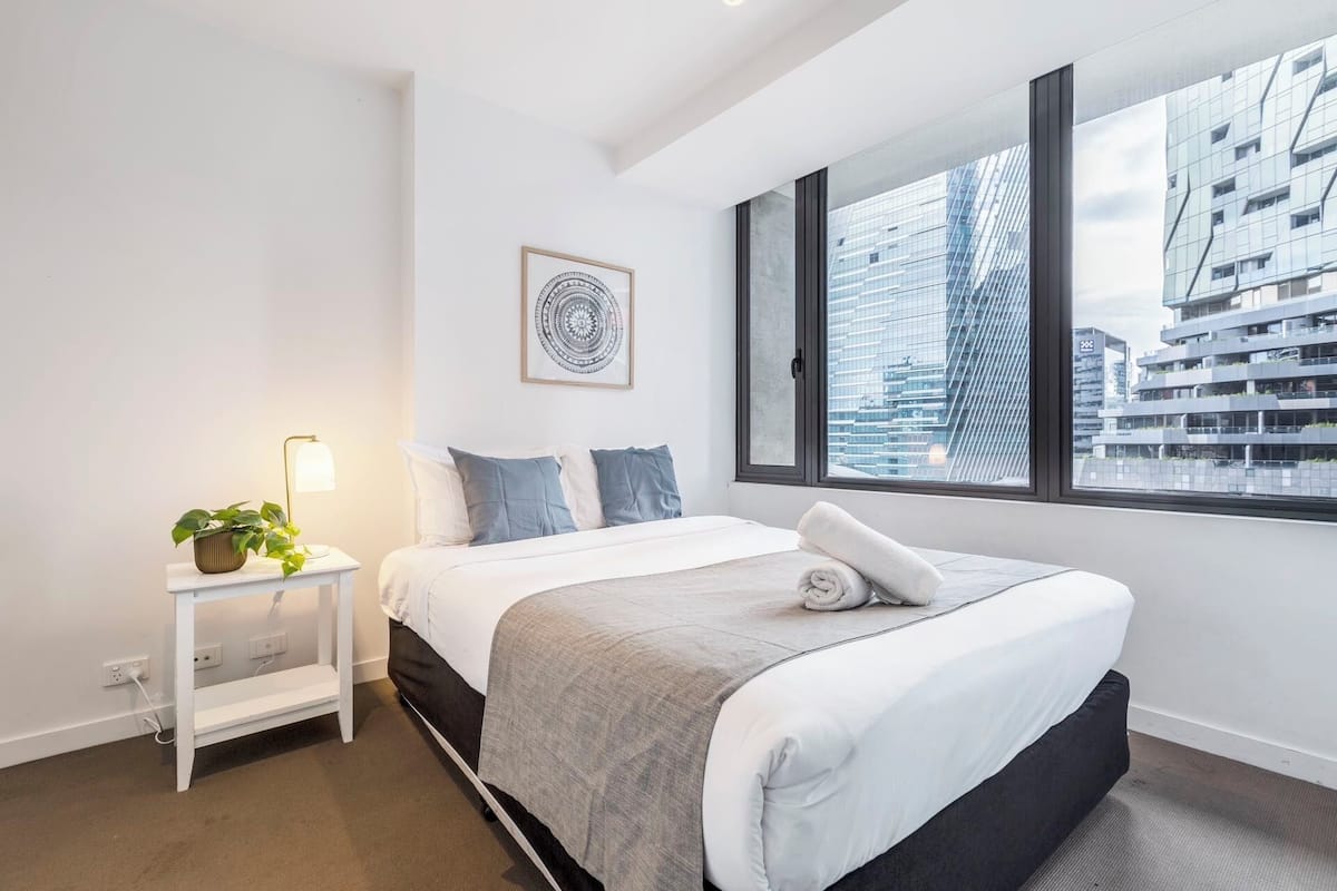A modern hotel room with a large bed, a bedside table with a lamp, a potted plant, and a view of a cityscape through a large window.