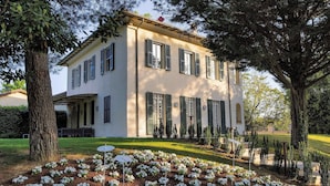 Un bâtiment à deux étages, doté d’une façade symétrique, de grandes fenêtres avec volets et d’un jardin bien entretenu agrémenté de fleurs blanches.
