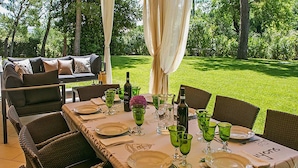 Un espace de restauration en plein air avec une table dressée pour six personnes, entourée de chaises en osier, d’un canapé et de rideaux.