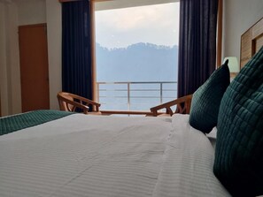 A hotel room with a large bed, two wooden chairs, a view of a lake and mountains, and a wooden headboard. - Amritara SunInn, Kedarnath (Ukhimath)