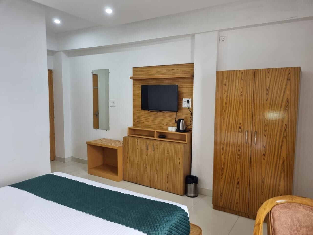 Habitación de hotel con una cama, un armario de madera, un televisor fijado a un panel de madera, un pequeño banco de madera y un cubo de basura.