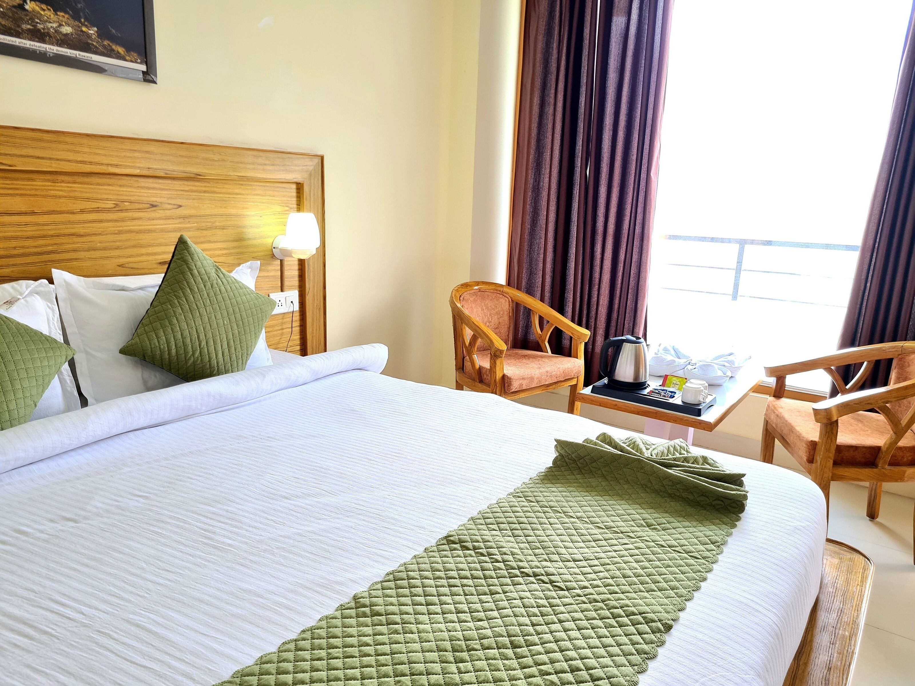 Una habitación de hotel con una cama grande, cabecera de madera, mesita de noche con lámpara, dos sillas, una pequeña mesa con hervidor y una ventana con cortinas.
