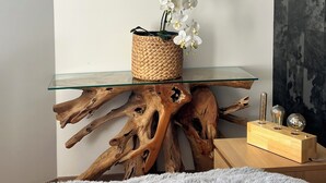 Une table à plateau en verre surmontée d’un panier tressé contenant des orchidées blanches, un bloc de bois sur lequel sont disposés des objets en verre, et une grande pièce de bois flotté servant de base.