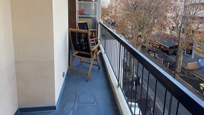 Ein Balkon mit Stuhl und Tisch, Blick auf eine Straße mit Gebäuden und Bäumen, und ein klarer Himmel.