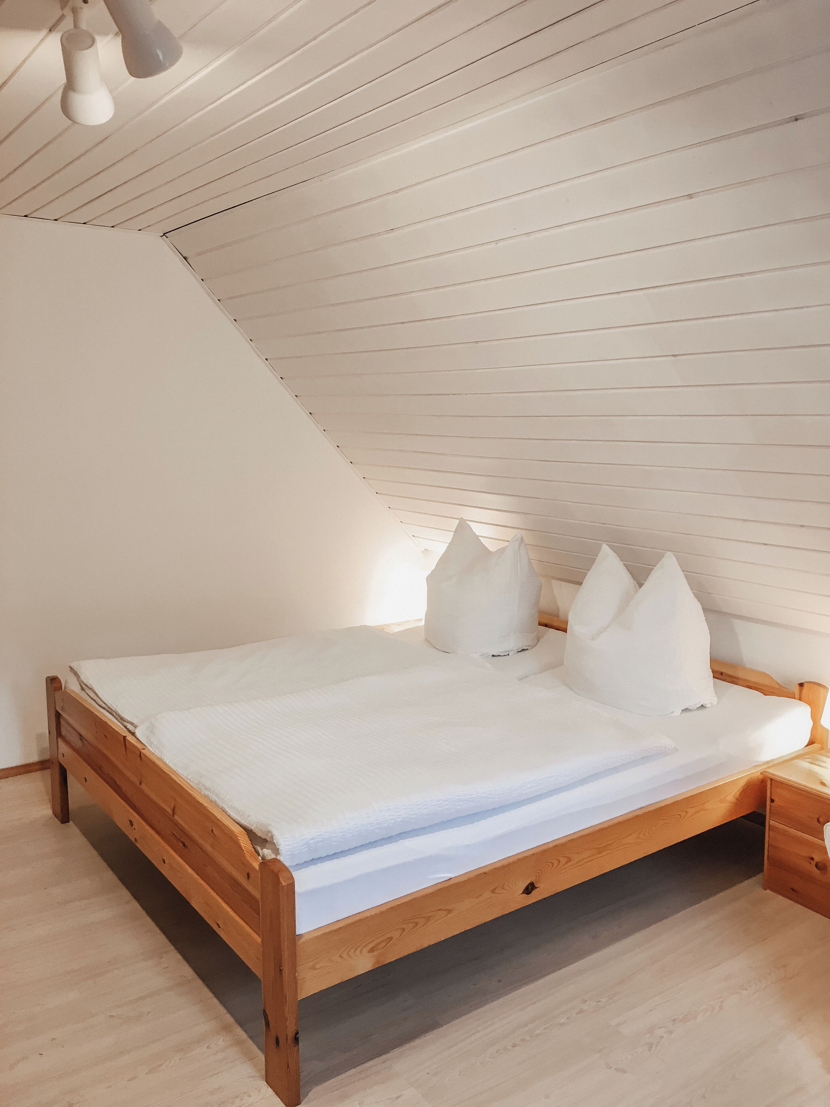 Une chambre avec un plafond en pente, un cadre de lit en bois, du linge de lit blanc et une table de chevet.