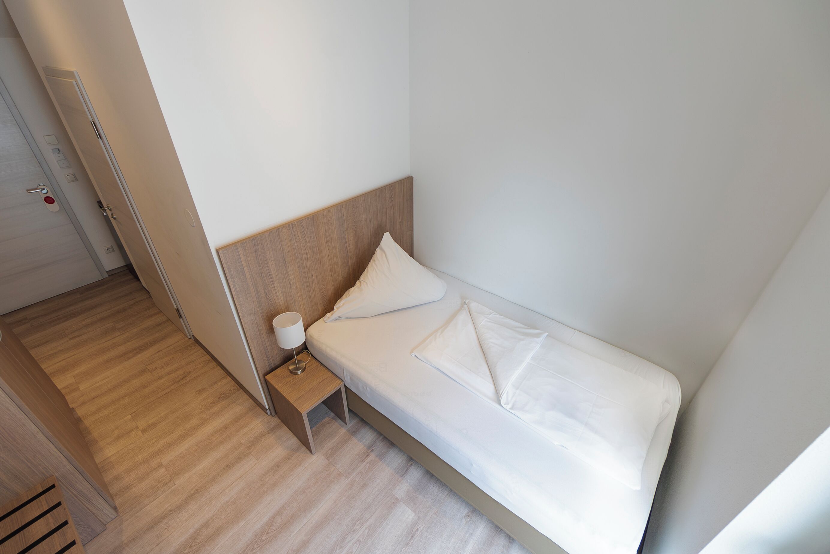 A single bed with a wooden headboard, a bedside table with a lamp, and a wooden floor.