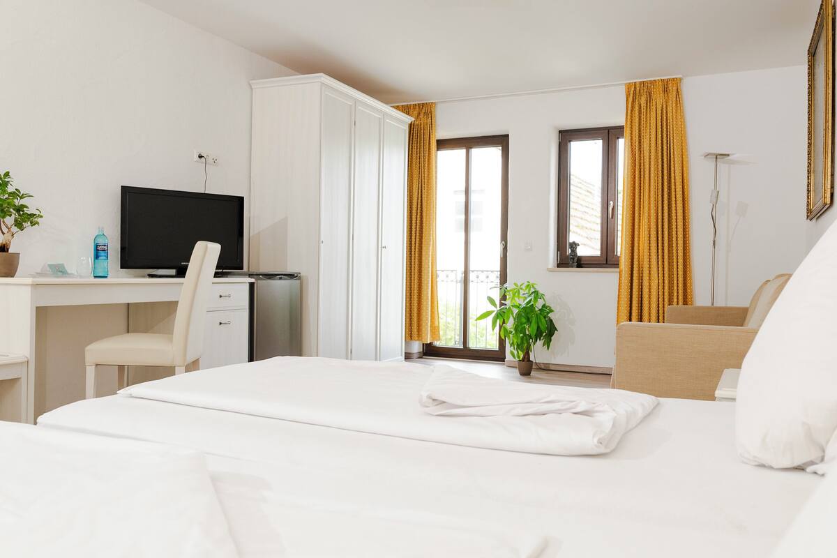 A hotel room with a bed, a desk with a TV, a chair, a potted plant, and a window with curtains.