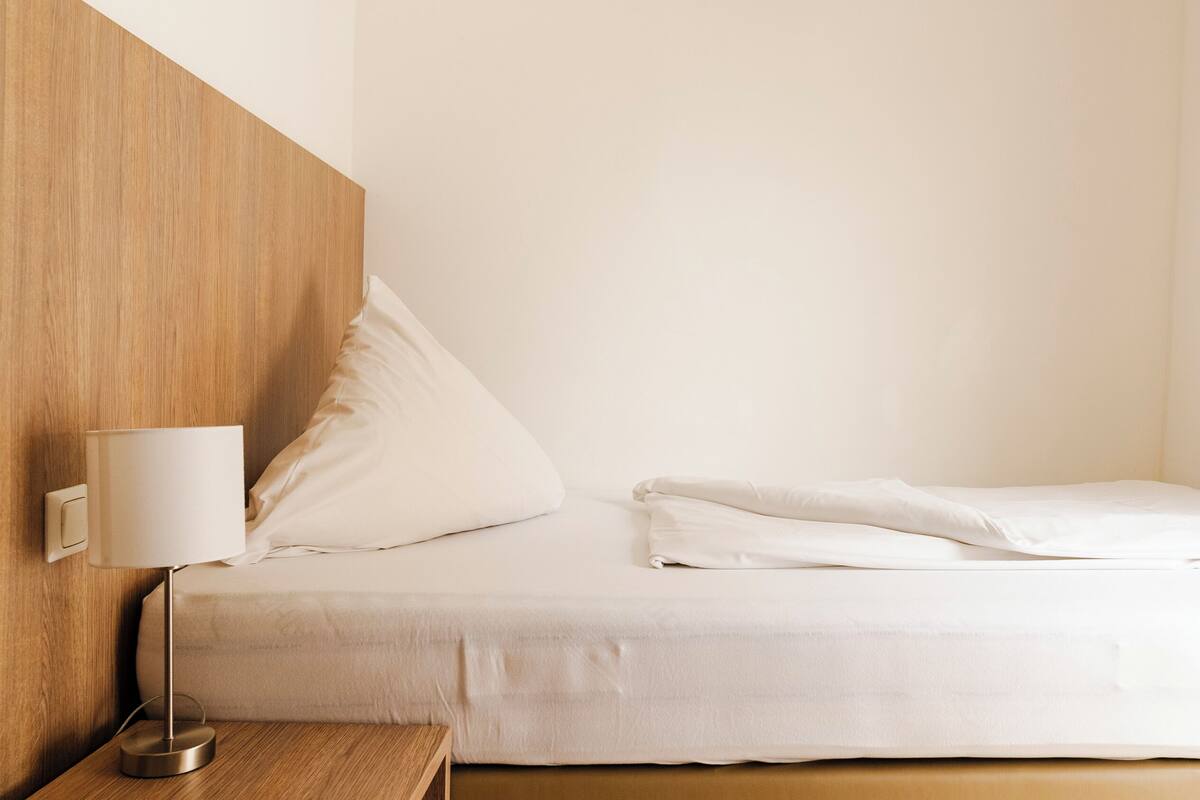 A neatly made bed with white linens, a bedside table with a lamp, and a wooden headboard.