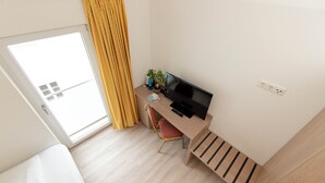 A small, modern room with a wooden desk, a TV, a chair, a potted plant, and a window with yellow curtains. - Henrys Sleeping Buttenwiesen (Buttenwiesen)