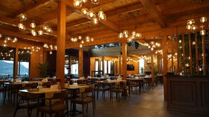 Un restaurante espacioso con numerosas mesas y sillas, vigas de madera en el techo, y lámparas esféricas colgantes.