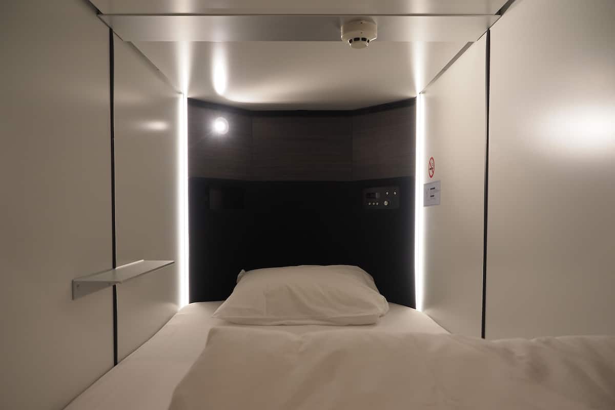 A compact, modern hotel room with a single bed, a control panel, and a ceiling-mounted light fixture.