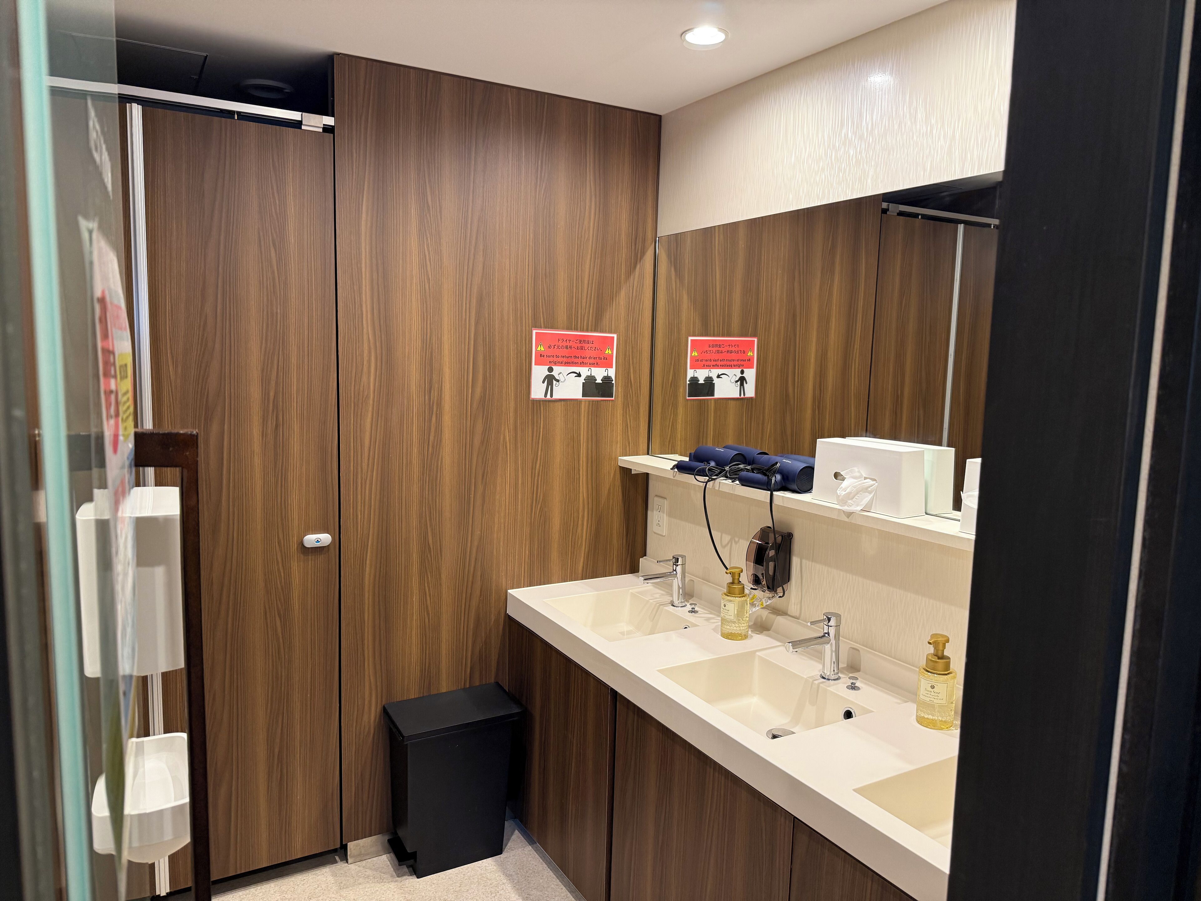 A modern bathroom with wooden paneling, a large mirror, two sinks, and a black trash bin.