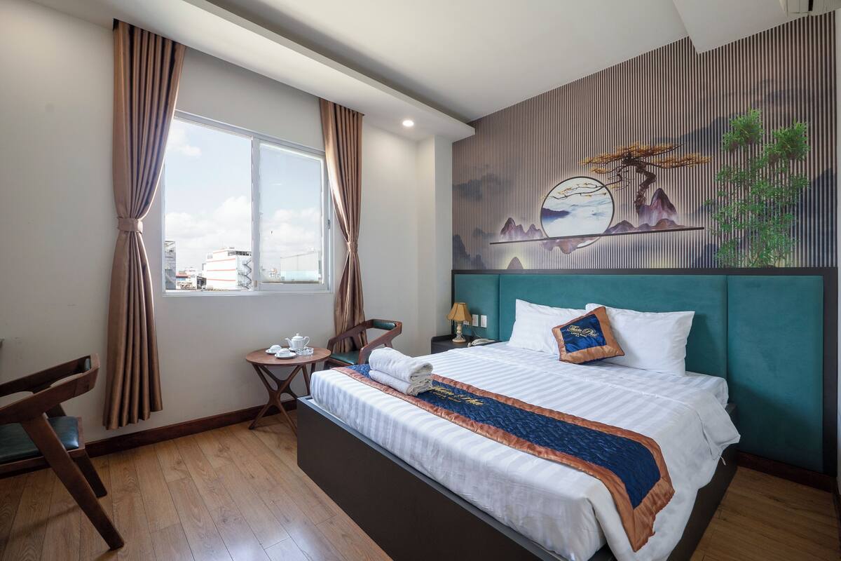 Une chambre d’hôtel avec un grand lit, une petite table avec une lampe, une chaise et une fenêtre avec des rideaux.
