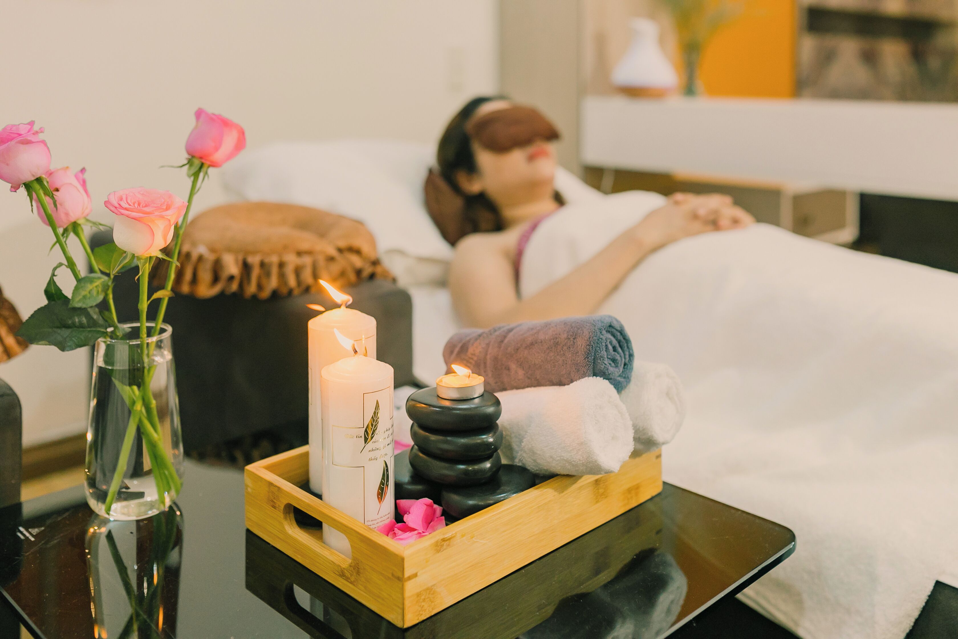 Une chambre à coucher avec une table de chevet sur laquelle se trouvent une bougie, un vase de roses et un plateau avec des serviettes et des pierres.
