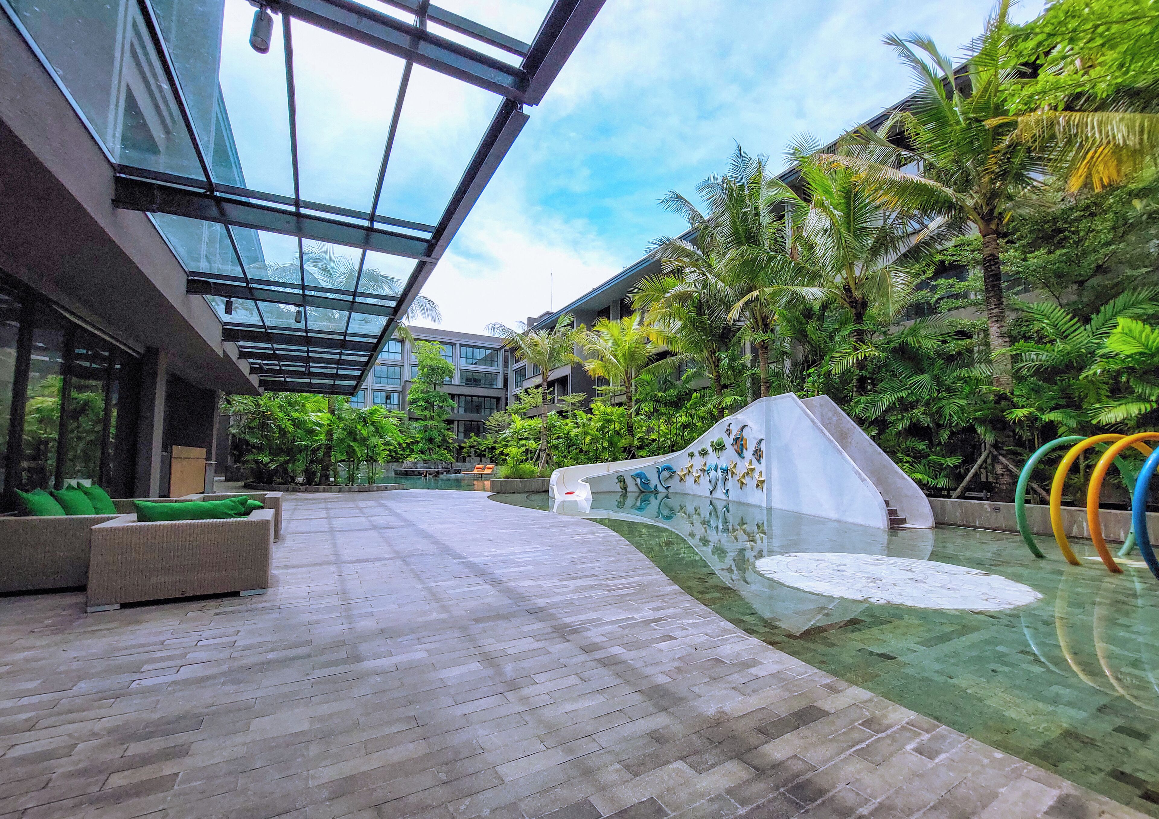 A modern indoor pool area with a glass ceiling, lounge seating, and tropical plants.