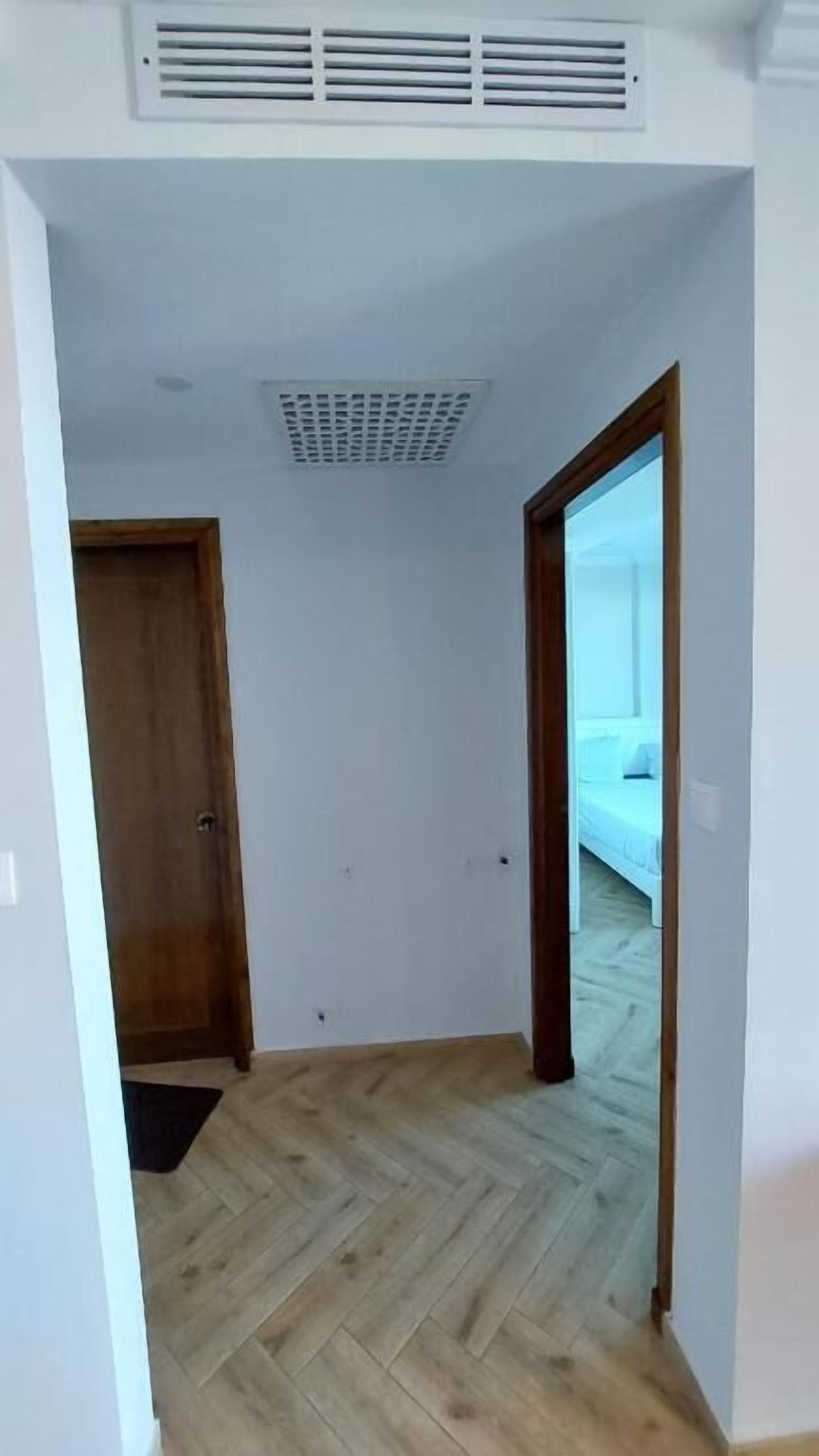 A room with a herringbone-patterned wooden floor, white walls, a wooden door, and a large window with a view of a bed.