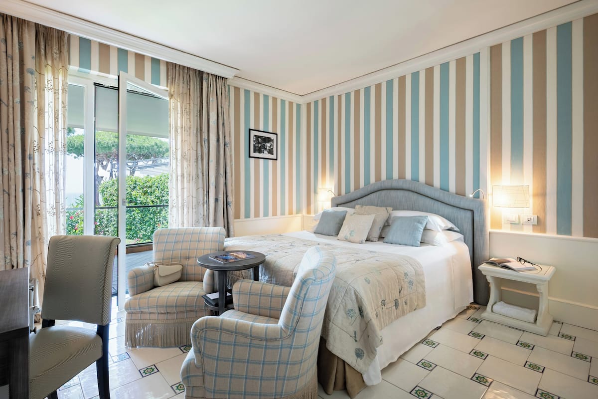 Habitación de hotel con cama, dos sillas, una mesita, una mesilla de noche y vistas al exterior con vegetación.