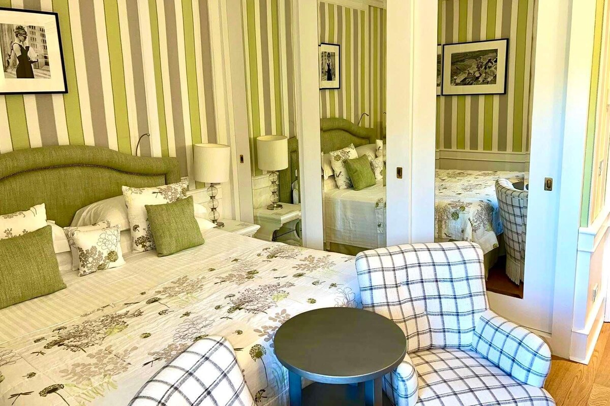 Una habitación con una cama grande, una silla, una mesita y un espejo.