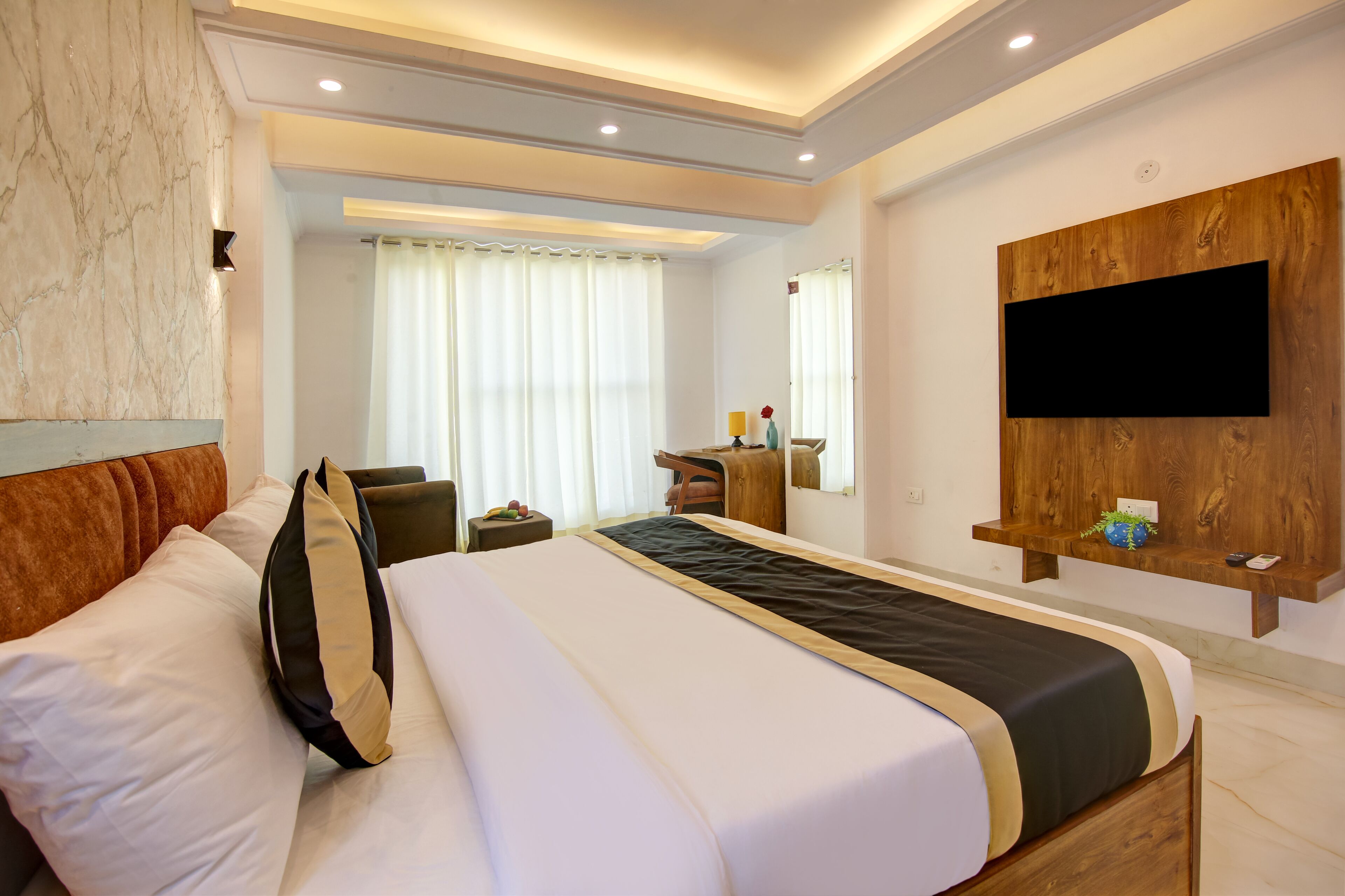 Quarto de hotel com uma cama grande, cabeceira de madeira, TV de tela plana fixada em um painel de madeira, pequena escrivaninha com cadeira e uma janela ampla com cortinas.