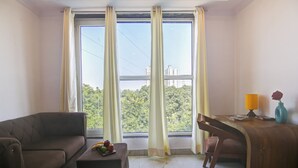 A room with a sofa, a small table with fruits, a desk with a lamp, and a view of greenery and buildings through large windows. - Hotel Emporio Grand (Ghaziabad)
