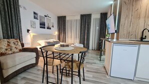 A modern living room with a sofa, a round wooden table with chairs, a small kitchen area with a refrigerator, and a TV on a wooden shelf. - Monfort A (Bansko)