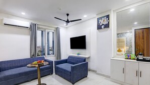 A modern living room with a blue sofa, a small round table with fruits, a ceiling fan, a flat-screen TV, and a kitchen area with cabinets and a countertop.