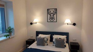 Un dormitorio con una cama, dos mesitas de noche, una ventana con planta y lámparas de pared.