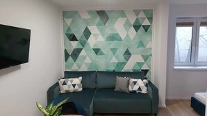 Un salón con un sofá verde, un pared con estampado geométrico y un televisor de pantalla plana montado en la pared.