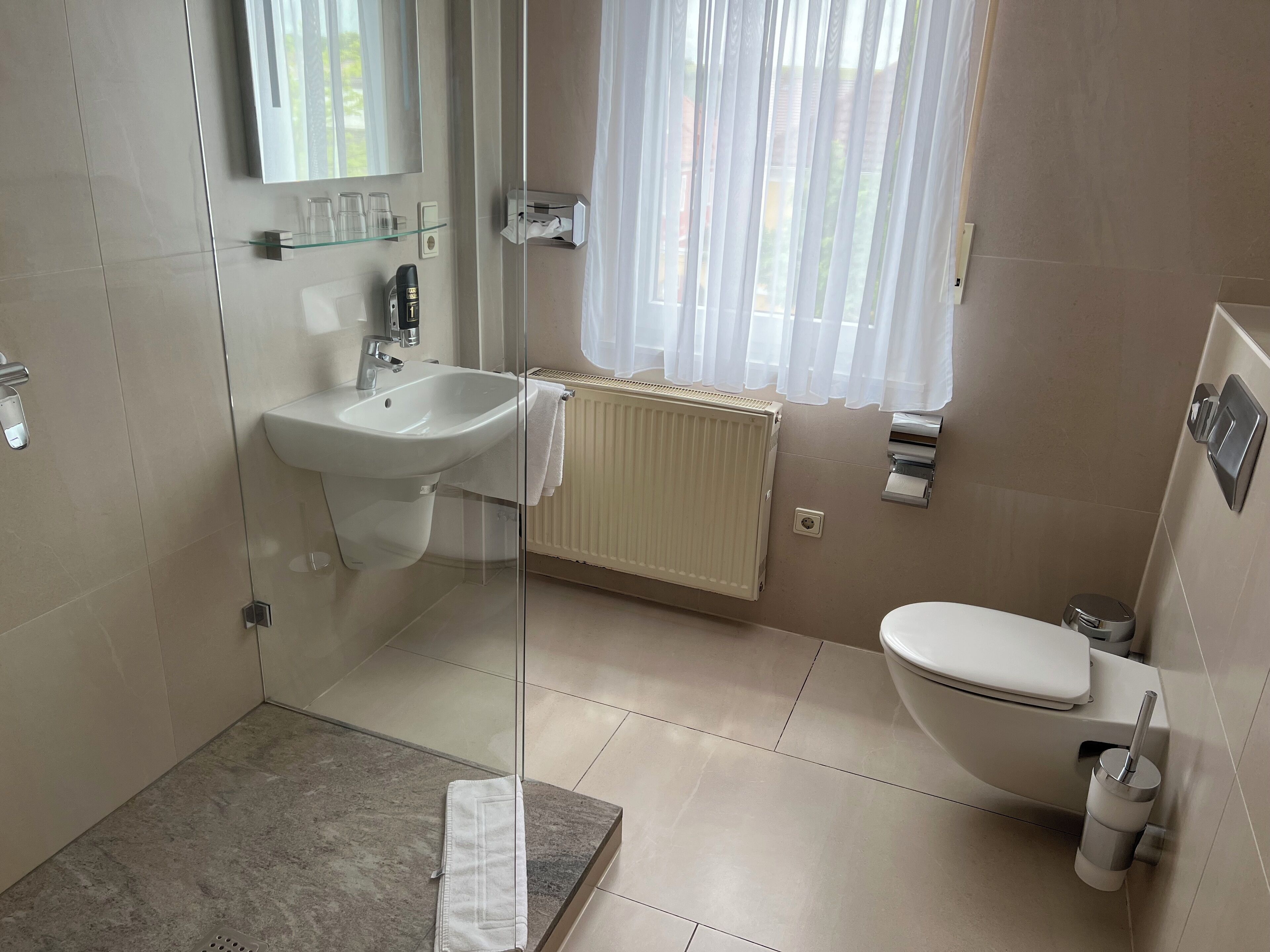 Ein modernes Badezimmer mit Duschkabine aus Glas, einem Waschbecken, einer Toilette und einem Fenster mit transparenten Vorhängen.