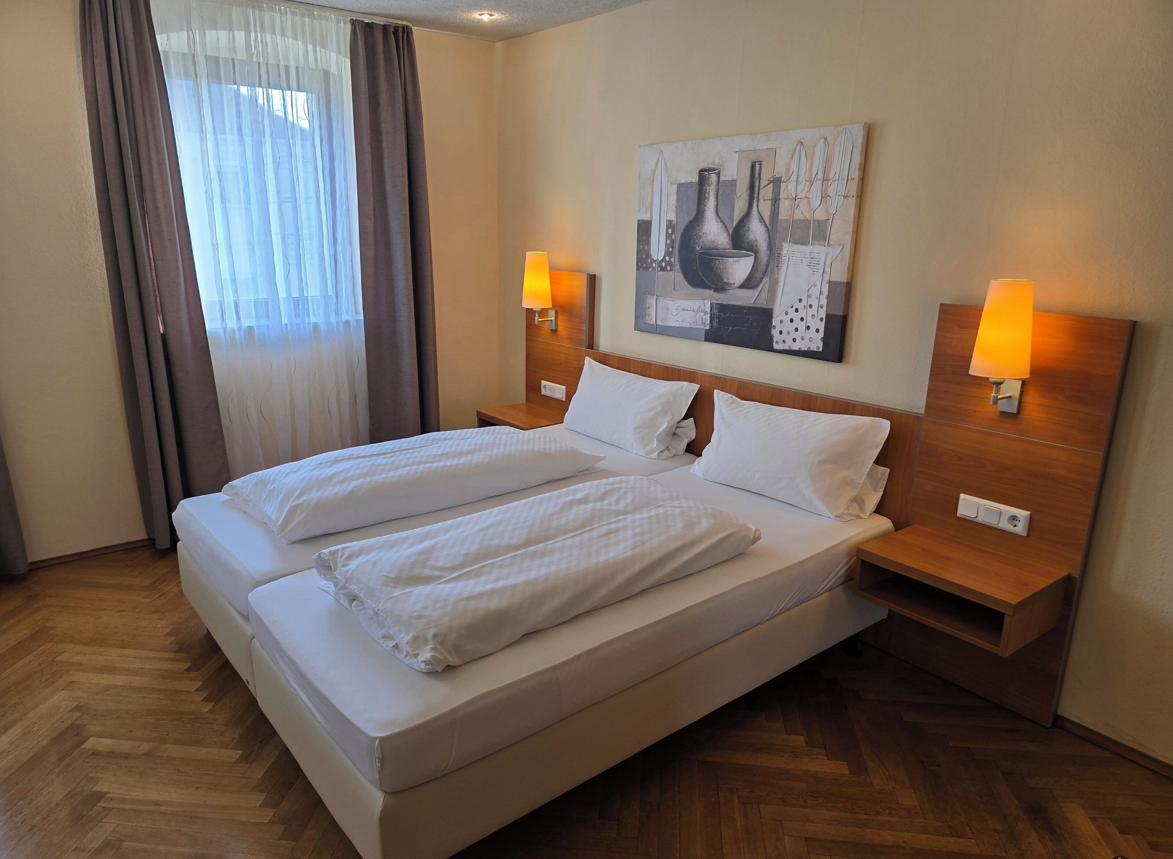 Ein Doppelbett mit weißen Leinen, zwei Nachttische mit Lampen, ein Gemälde an der Wand und ein Fenster mit Vorhängen.