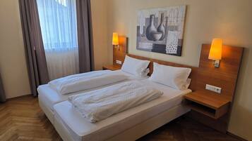 A double bed with white linens, two bedside tables with lamps, a painting on the wall, and a window with curtains.