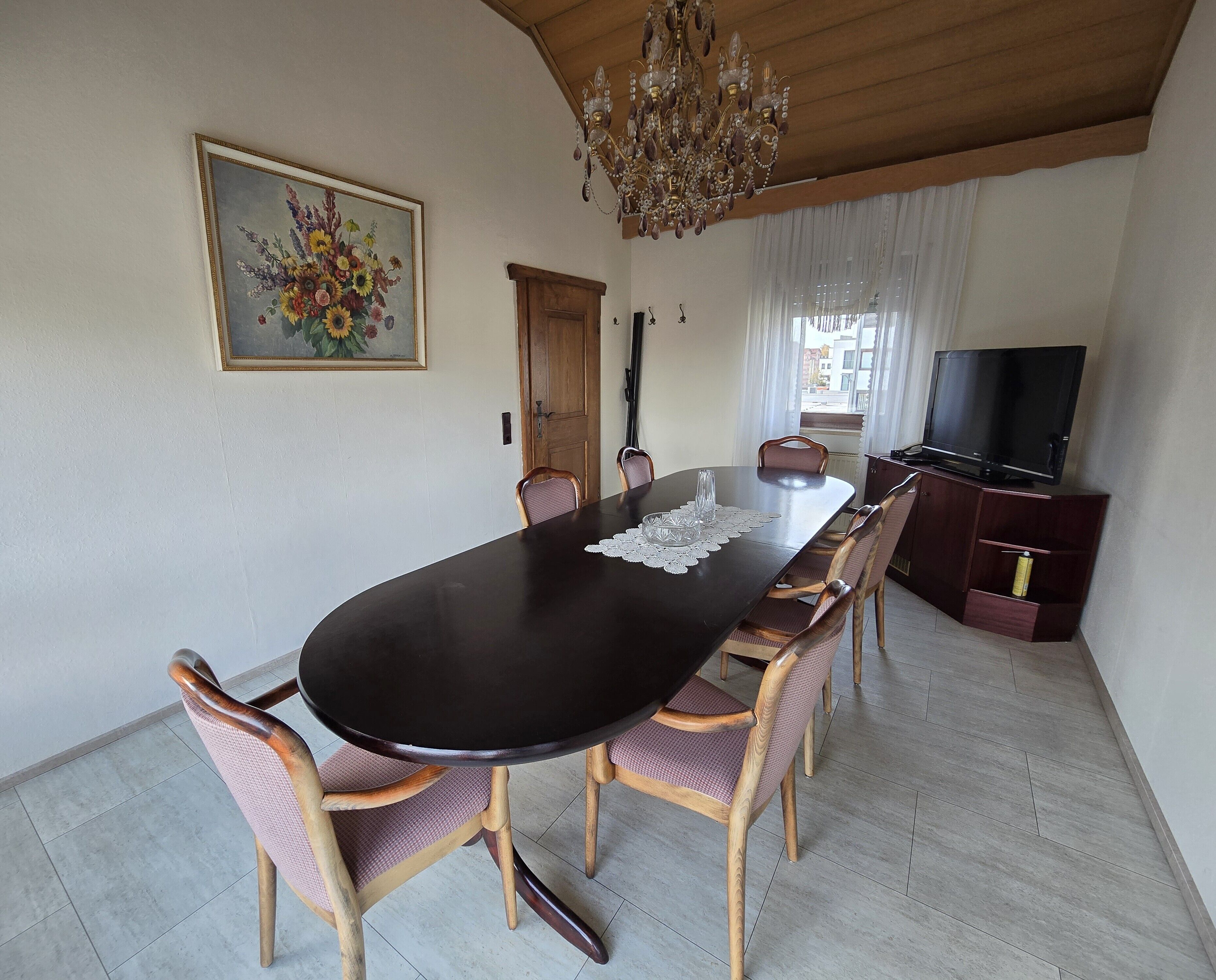 A dining area with a large table, chairs, a chandelier, a painting, and a television.