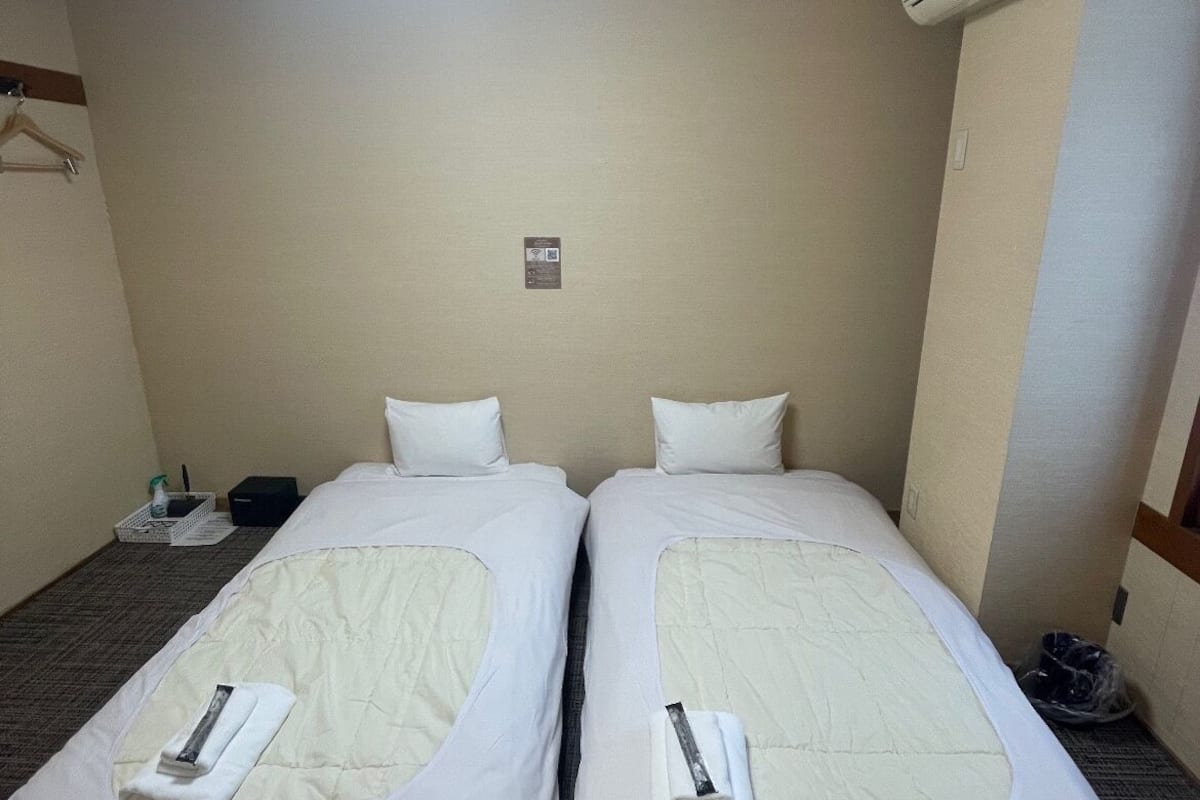 Ett hotellrum med två separata sängar, ett litet bord med en flaska och en näsdukask, samt en luftkonditioneringsanläggning på väggen.