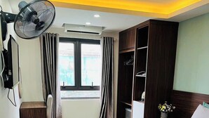 A hotel room with a large bed, a wardrobe, a desk, a chair, a standing fan, and a window with curtains. - Green Hotel Apartment HN- by Bay Luxury (Hanoi)