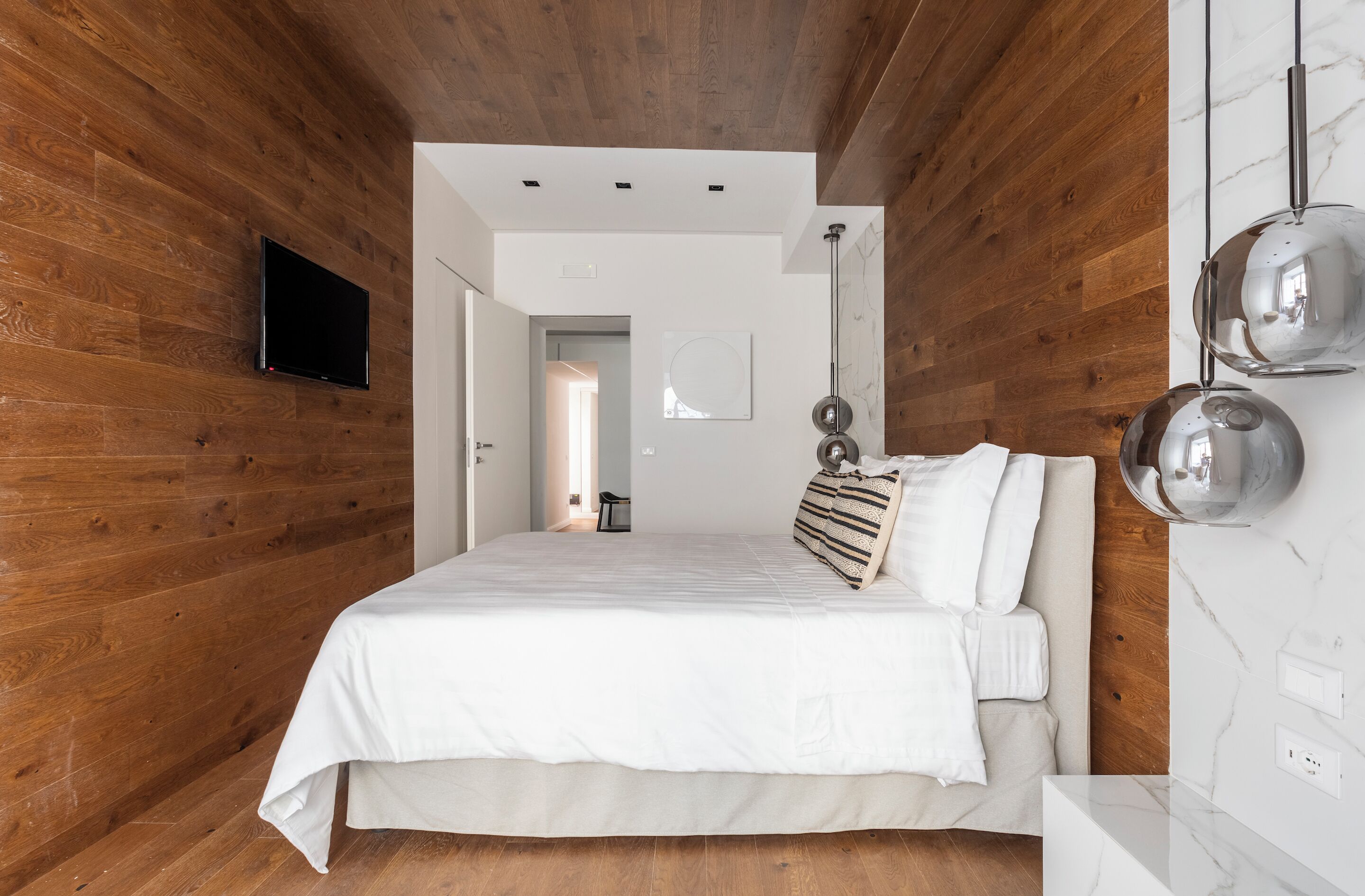 A bedroom with a wooden wall, a bed with white bedding, a TV mounted on the wall, and a marble wall with a light fixture.