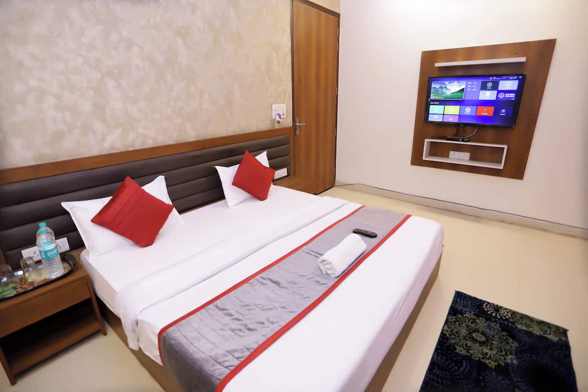 Habitación de hotel con cama, televisión montada en la pared y mesita de noche con una botella y vasos.