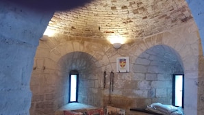 Una habitación de paredes de piedra con una cama, almohadas con diseños históricos, un escudo en la pared y una ventana pequeña.