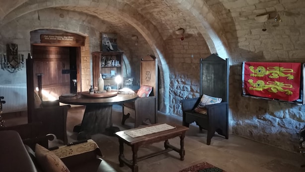 Una habitación rústica con techo de piedra, una mesa de madera, un sofá y una bandera roja con un emblema de león en la pared.