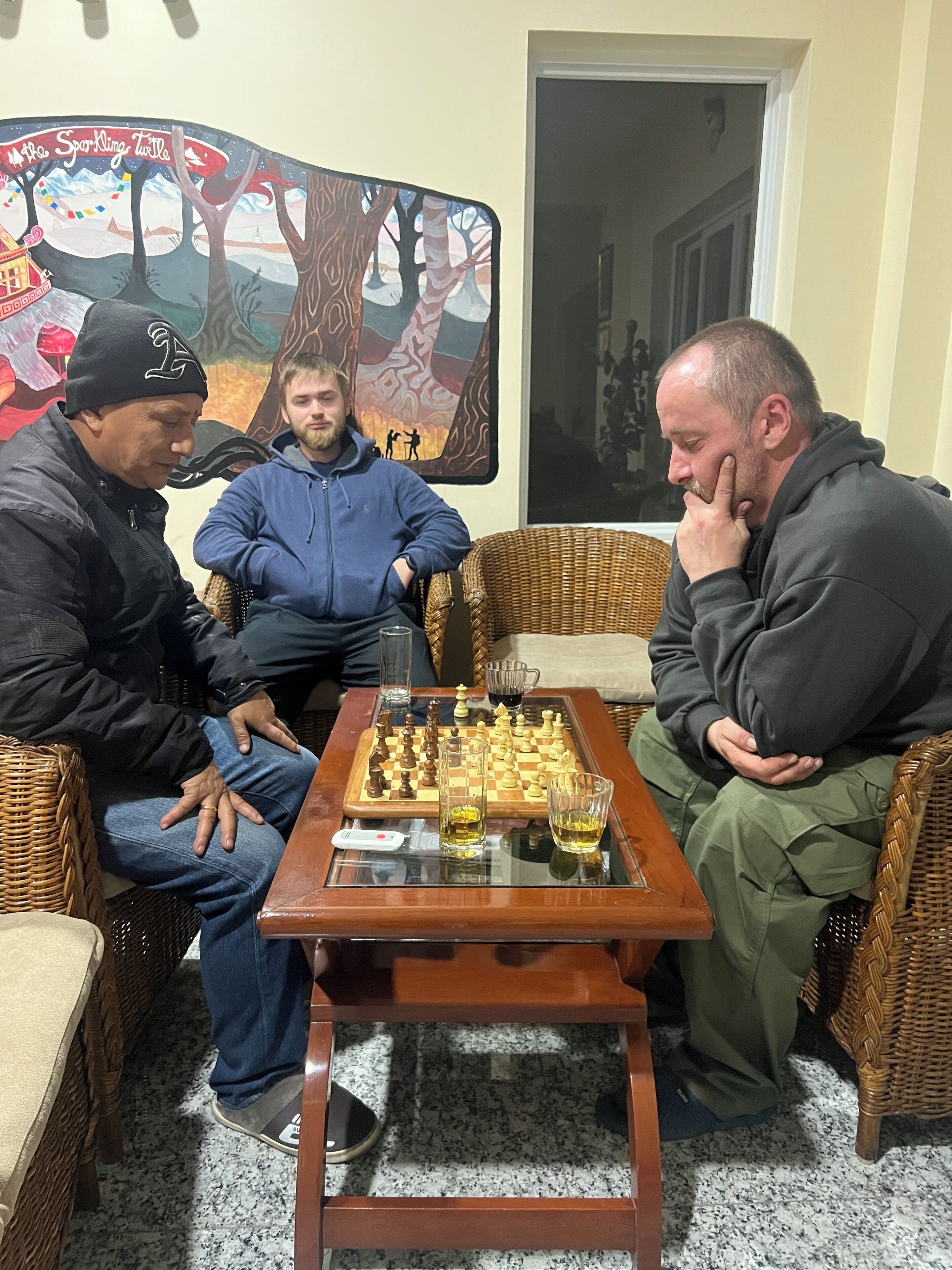 Three people playing chess on a table with glasses of alcohol, in a room with wicker furniture and a painting on the wall.