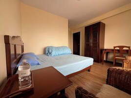 Habitación de hotel con cama, mesita de noche de madera con una botella de agua, una pequeña mesa, una silla de mimbre y un armario.