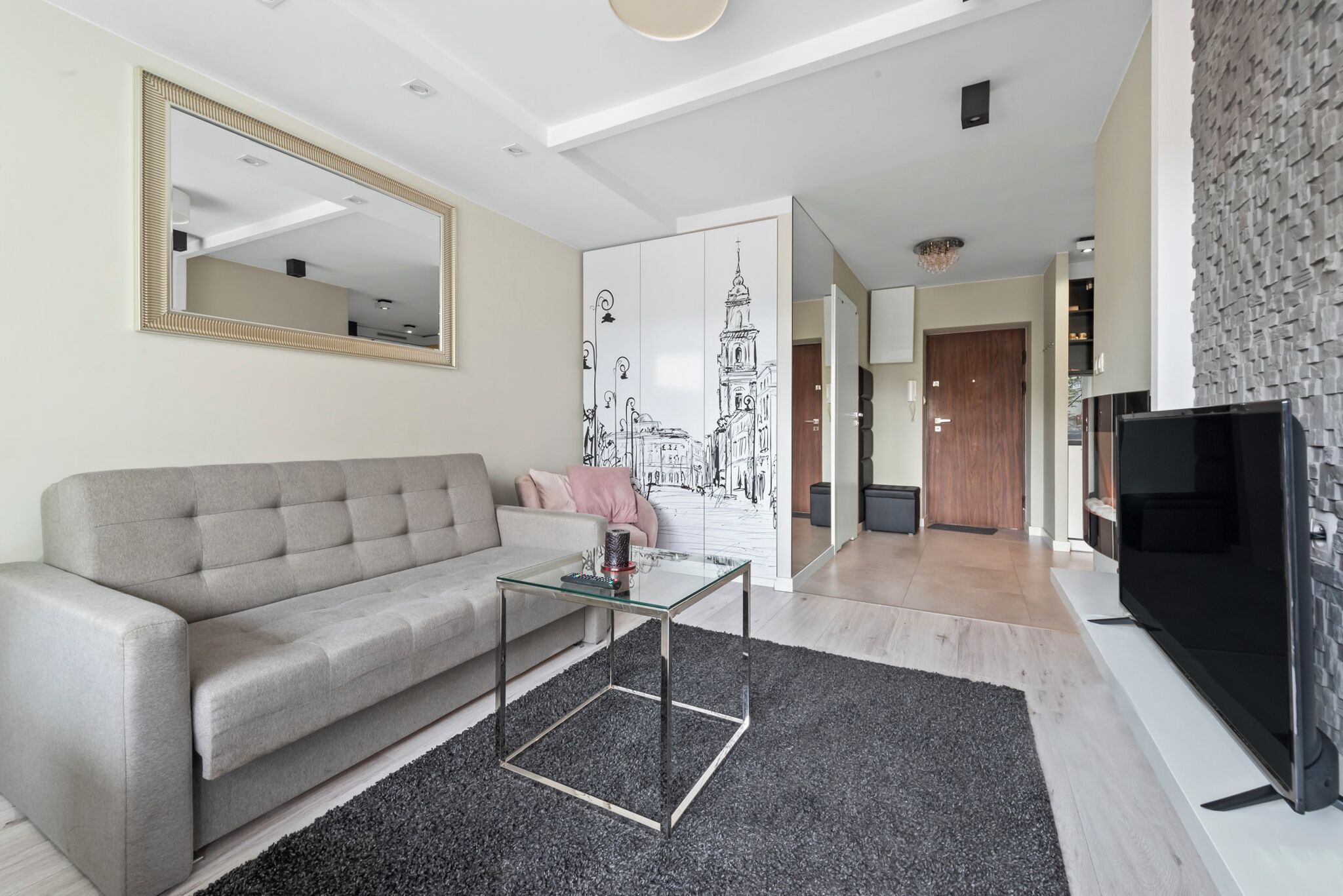 Un salon moderne avec un canapé gris, une table basse en verre, un téléviseur noir et un grand miroir.