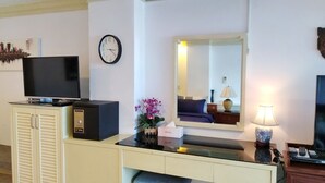 A hotel room with a television, a clock, a mirror, a desk with a lamp, and a cabinet. - Spacieux Studio, Tres Belle vue mer sur la Baie de Patong (Tambon Patong)
