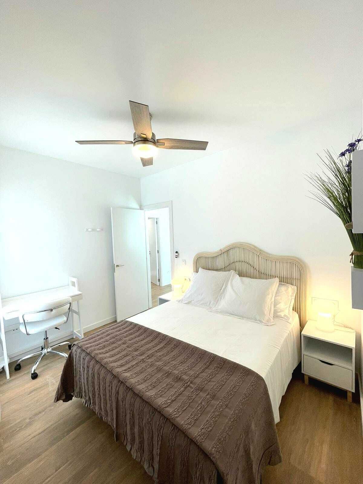 A bedroom with a bed, a desk, a chair, a nightstand, a lamp, and a ceiling fan.