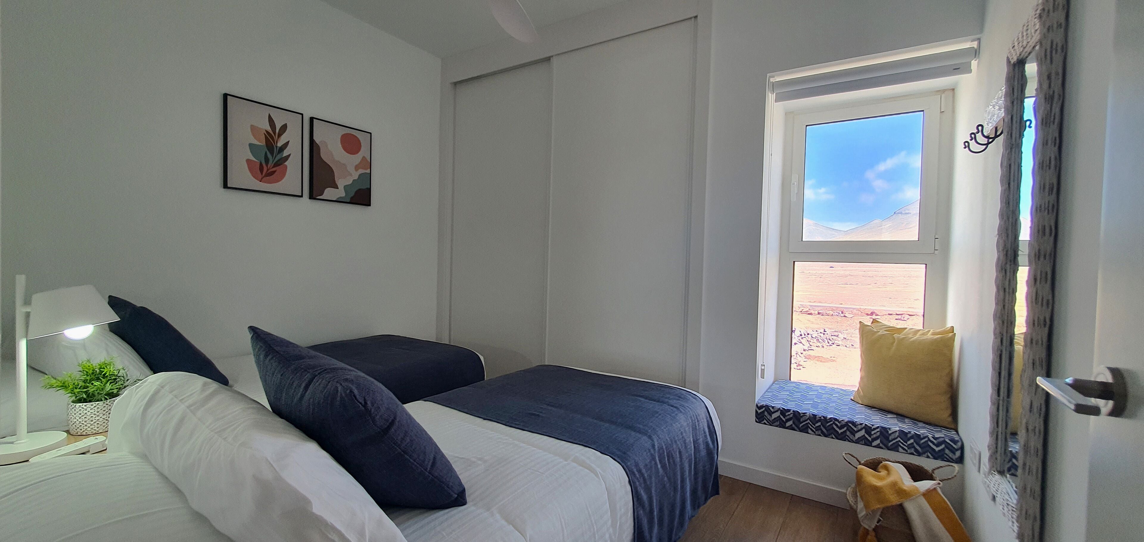 A bedroom with two beds, a window seat with a view, and a small table with a lamp and plant.