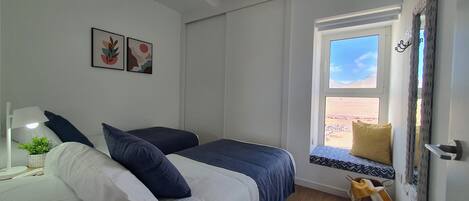 A bedroom with two beds, a window seat with a view, and a small table with a lamp and plant.